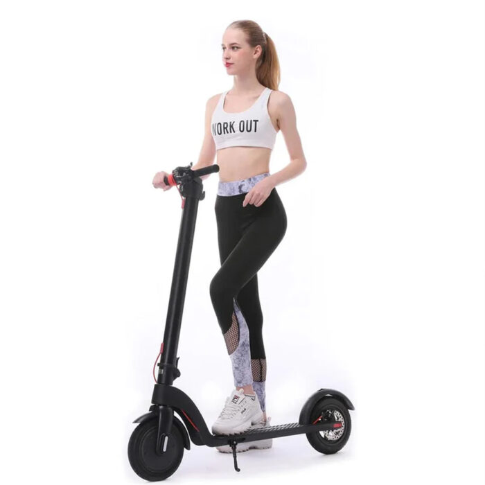 A woman in workout clothes stands next to an X7 500w Foldable E-Scooter With Replaceable/Detachable Lithium Battery, 8.5-inch pneumatic tires (Black), wearing a white WORK OUT sports bra, black mesh-detailed leggings, and white sneakers.